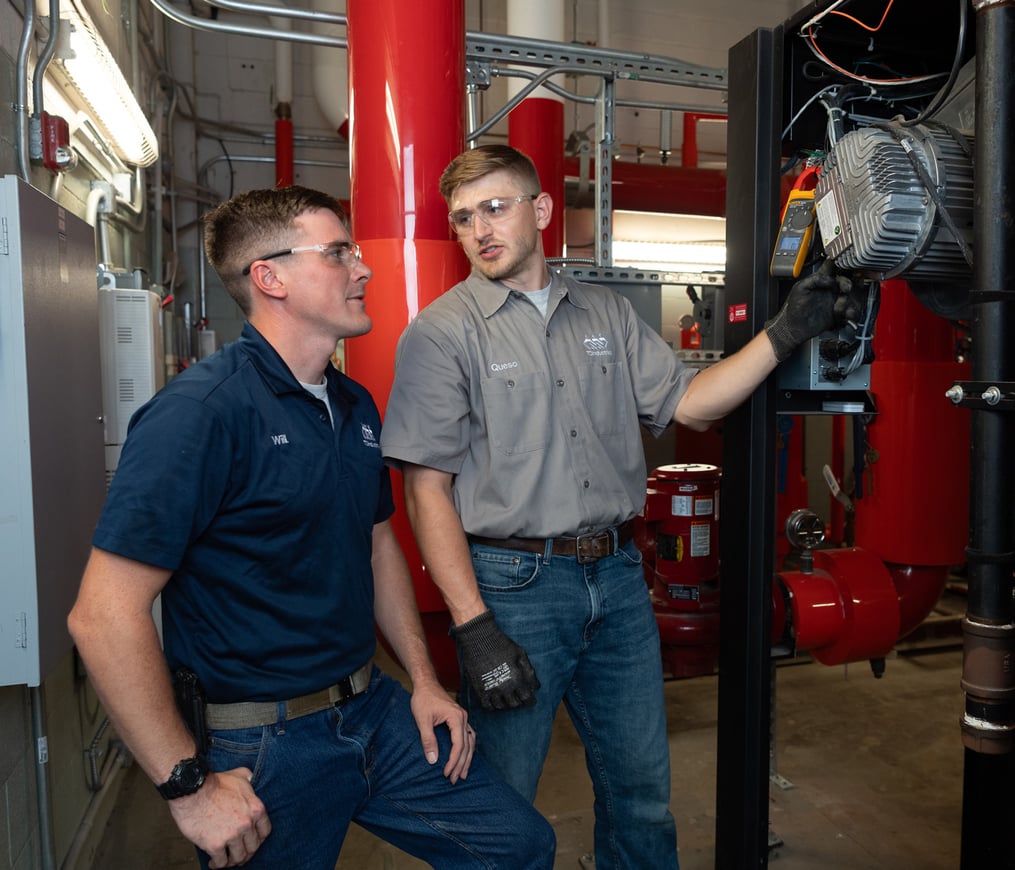 Two TD plumbers discuss turnkey plumbing services and compliance in a school mechanical room